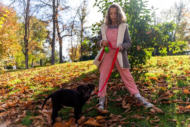 A dog and its owner in a park. The dog is wearing a long leash and is a few feet away from the owner. The owner is kneeling and smiling at the dog, holding a treat in one hand and the leash in the other.