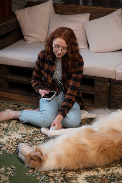A person sitting on the floor, interacting with their dog, who is lying down calmly. The person is petting the dog gently, creating a relaxed and affectionate atmosphere. The background is a cozy living room setting.